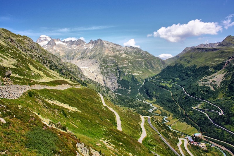 Winding pass road between green Alpine slopes, with glaciers and peaks behind